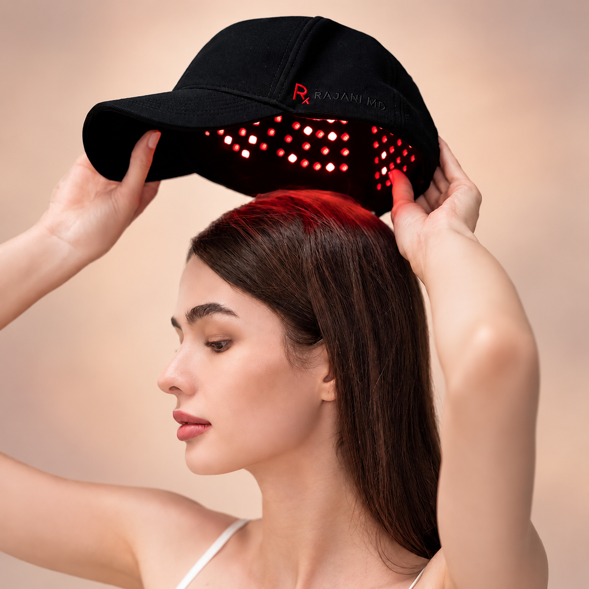 A woman puts on a Rajani MD PlasmaGLO Hair+ Cap with the laser and LED red light therapy panels illuminated.