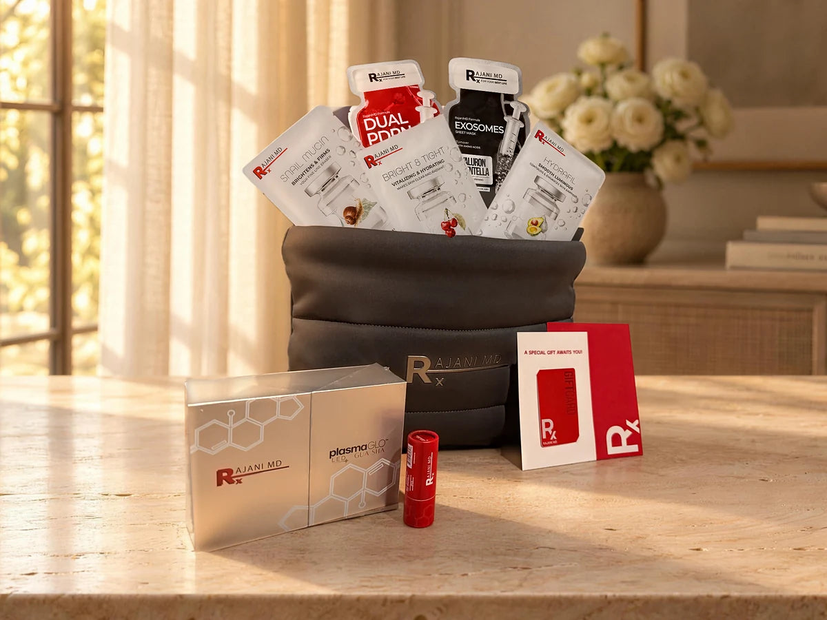 The RajaniMD guest bag with five sheet masks, the plasmaGLO LED Gua Sha, a lip balm and the red gift card, on a stone table in soft window light