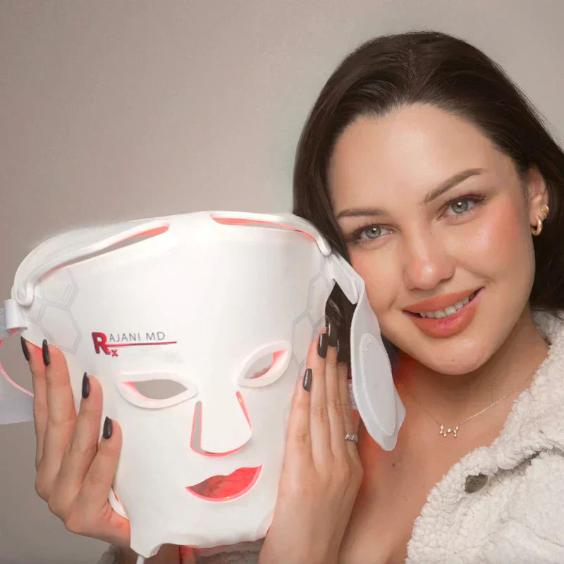 A smiling woman holds a glowing Rajani MD red light therapy face mask beside her face.